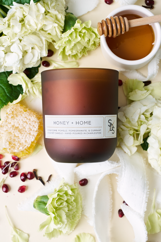 The Honey + Home Signature Candle, hand-poured in Charleston, is brown and displayed with white flowers, a honey dipper in honey, honeycomb, pomegranate seeds, and green leaves on a light background.