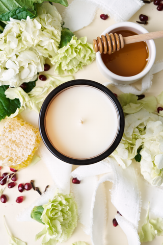 The Honey + Home Signature Candle, hand-poured in Charleston, glows in a black jar amid white flowers, honeycomb, a honey dipper with honey, pomegranate seeds, and soft white fabric on a light surface.
