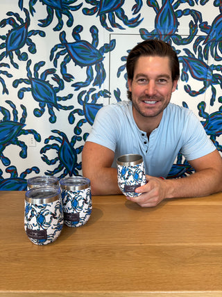 A man in a light blue shirt holds a Crab Craze Shades of Blue Wine Tumbler and stands behind a table with three similar cups. The wall adorned with a blue crab pattern complements the tumblers design, as he smiles at the camera.