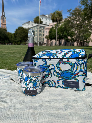A picnic on grass features a colorful Sewing Down South pattern cooler bag and two Crab Craze Shades of Blue Wine Tumblers with a blue crab design. A champagne bottle sits behind them, set against a charming backdrop of a church and trees.