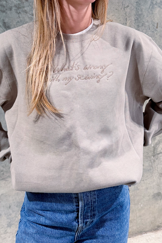 Wearing the WWWMS Embroidered Script Crewneck Sweatshirt in beige, which reads "Whats wrong with my sewing?", a person with long blonde hair and blue jeans stands against a concrete wall.