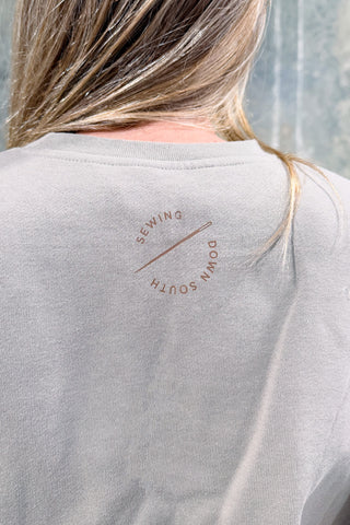 A person with blonde hair wears a light gray WWWMS Embroidered Script Crewneck Sweatshirt featuring a circular "SEWING // DOWN SOUTH" logo on the upper back.