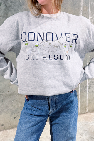 A person wearing a gray Conover Ski Resort Crewneck Sweatshirt with a mountain and ski lift graphic stands against a textured concrete wall.