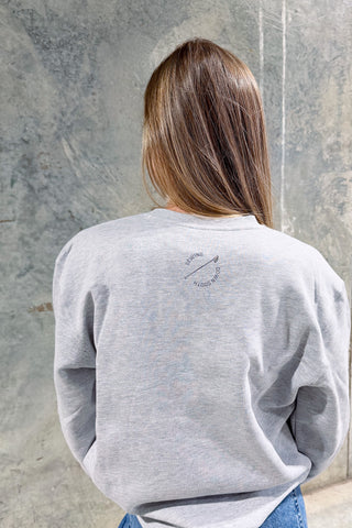 A person with long brown hair wears a light gray Conover Ski Resort Crewneck Sweatshirt, standing with their back to the camera against a textured gray wall. Small circular text on the upper back adds an apres-ski vibe.