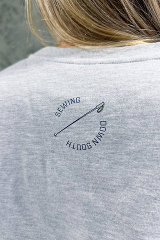 Close-up of the back of a gray Conover Ski Resort Crewneck Sweatshirt featuring a small needle and thread graphic with "SEWING DOWN SOUTH" curved around it. Ideal for mountain getaways. The wearer’s hair is partially visible.