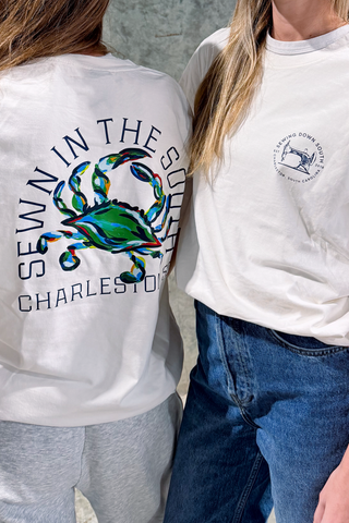 Two people stand side by side in white tees with coastal flair. One wears the Crab Craze Tee, featuring "Sewn in the South Charleston SC." The other sports a retro unisex shirt with a small circular chest logo.