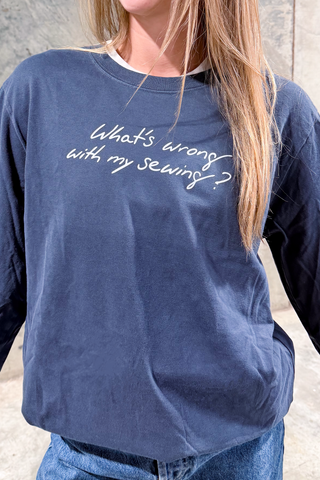A person with long light brown hair stands against a light stone background, wearing the WWWMS Script Long Sleeve Tee featuring white "What's wrong with my sewing?" text in Feather: Craig's Handwriting.
