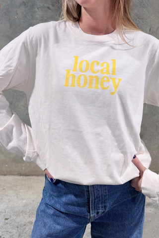 Wearing the Local Honey Long Sleeve Tee in soft ring-spun cotton with yellow front lettering, this unisex relaxed fit top pairs easily with blue jeans, shown here against a neutral gray backdrop.