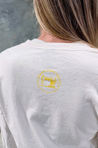 A person with long, light brown hair wears the Local Honey Long Sleeve Tee, made from 100% ring-spun cotton, featuring a yellow sewing machine and circular text graphic on the back. The softly blurred gray background adds Southern charm.