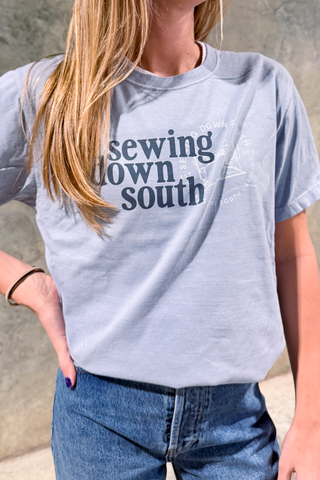 A person models the SDS Brand Logo Tee in light blue with blue jeans, posing with one hand on their hip against a neutral backdrop.