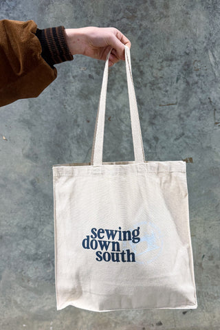 A person in a brown jacket holds the SDS Brand Logo Tote, a cotton bag featuring "Sewing Down South" in dark blue on the front, set against a gray concrete background.