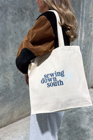 Carrying the beige SDS Brand Logo Tote printed with blue text, a person with long wavy hair, dressed in a brown and black jacket and light pants, stands by a concrete wall.