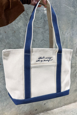 A hand holds the WWWMS Embroidered Script Canvas Tote, a cream heavyweight cotton bag with blue straps and trim. “What’s wrong with my sewing?” is embroidered on the accessories pocket, shown against a textured gray wall.
