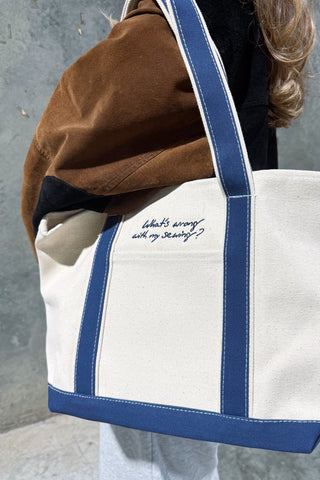 A person wearing a brown and black jacket carries the WWWMS Embroidered Script Canvas Tote, a cream bag with blue straps featuring the text, "What's wrong with my sewing?".