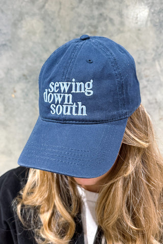 A person with long, wavy blonde hair wears an SDS Embroidered Logo Hat—a blue premium cotton twill cap with “sewing down south” stitched in white and an adjustable buckle—as they look down.