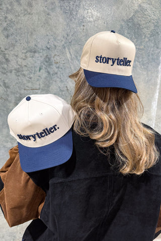 A person with long, wavy blonde hair wears the cream and navy Storyteller 3D Embroidered Hat. A second identical Bravocon exclusive cap sits on their shoulder against a neutral background.