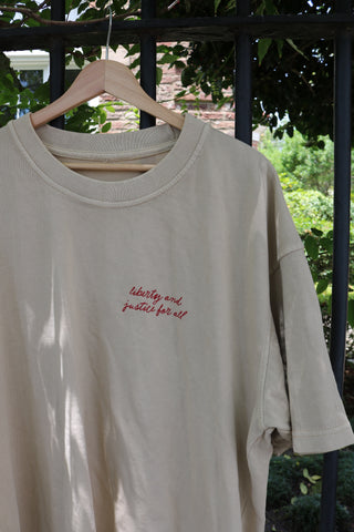 The Loyal Liberty Tee, part of the Americana Capsule Collection, is a beige heavyweight cotton shirt with red "liberty and justice for all" embroidery, shown on a wooden hanger outdoors against a black metal fence and green foliage.