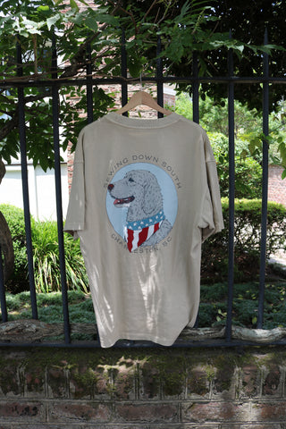 The Loyal Liberty Tee, a beige shirt from the Americana Capsule Collection, hangs on a black metal fence. It features a dog in an American flag bandana and “SEWING DOWN SOUTH CHARLESTON, SC” text, with greenery in the background.
