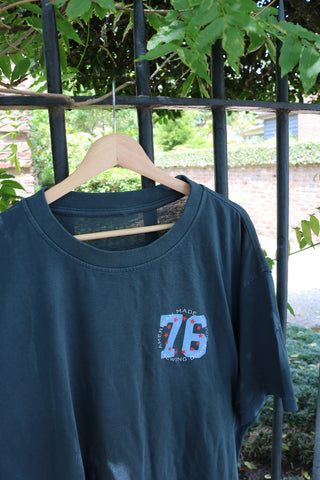 The 1776 Tee, featuring "76" and "Made for Moving" on the chest, hangs on a wooden hanger by a metal fence with green leaves and a brick wall—part of our Americana Capsule Collection.