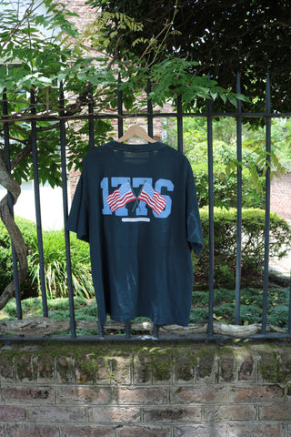 The 1776 Tee from the Americana Capsule Collection hangs on a black iron fence, showcasing “1776” above the Liberty Bell and two American flags, with greenery and a brick wall in the background for a historic touch.