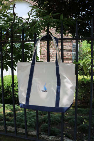 The Set Sail Embroidered Tote, a white cotton canvas bag with blue handles, trim, and a blue sailboat design on the front, hangs on a black fence with green foliage and a brick building in the background.