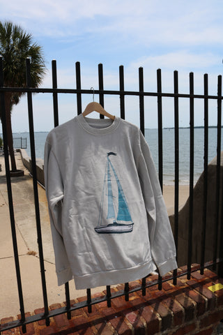 The Set Sail Crewneck Sweatshirt from the Americana capsule, in light gray with a sailboat graphic, hangs on a wooden hanger from a black metal fence. A palm tree and partly cloudy waterfront are visible in the background.