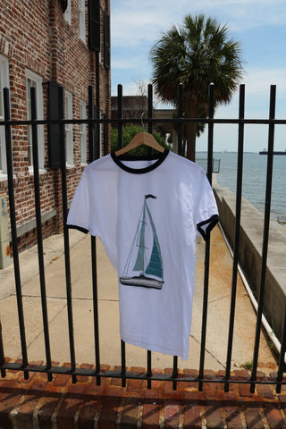 The Set Sail Ringer Tee, featuring a vintage sailboat graphic, hangs on a fence by a brick building with palm trees and a waterway in the background—part of our Americana Capsule Collection.