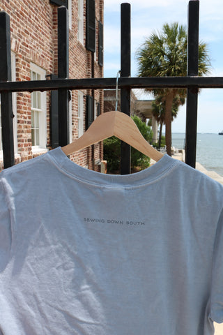A Set Sail Women's Tee in light blue hangs on a wooden hanger from a black iron fence. SEWING DOWN SOUTH is printed on the shirt, with a brick building, palm tree, and waterfront in the background for summer vibes.