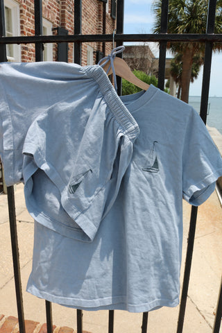 The Set Sail Women's Shorts in light blue, paired with a matching short-sleeved shirt featuring sailboat embroidery, hang on a wooden hanger on a black iron fence with water, palm trees, and buildings in the background.