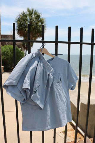 The Set Sail Women's Shorts from the Americana Capsule Collection, in light blue with sailboat embroidery, hang on a hanger from a black iron fence by the ocean, framed by a palm tree and sidewalk for a breezy summer vibe.