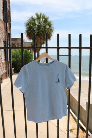 A Set Sail Women's Tee in light blue, featuring a small sailboat graphic, hangs on a wooden hanger from a black metal fence with a palm tree, sidewalk, and ocean visible in the sunny summer background.