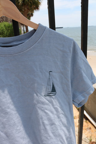 The Set Sail Women's Tee, in light blue with a sailboat chest design, hangs on a wooden hanger by metal bars. A sandy beach and ocean serve as the backdrop—ideal for effortless summer style.