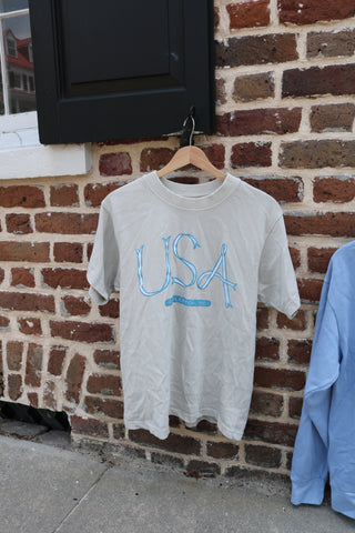 The USA Ribbon Tee from the Americana Capsule Collection hangs on a wooden hanger against a brick wall by a window. Made from heavyweight cotton, it features bold blue lettering for classic American style.