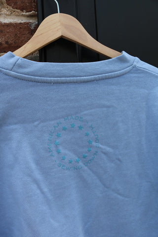 A light blue USA Ribbon Crewneck Sweatshirt hangs on a wooden hanger, featuring a circular teal logo with text and stars on the upper back, set against a dark door and brick wall.