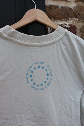 The USA Ribbon Tee, a heavyweight cream cotton shirt with a blue circular AMERICAN MADE HANDS SEWING DOWN logo and stars, hangs on a wooden hanger against a brick wall and black door. Part of the Americana Capsule Collection.
