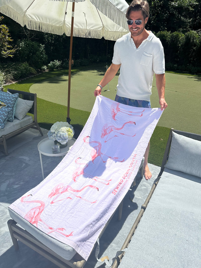 A smiling man in sunglasses and a white shirt lays the Flamingo Beach Towel on a lounge chair outdoors beside a small table with white flowers, near an umbrella and greenery.
