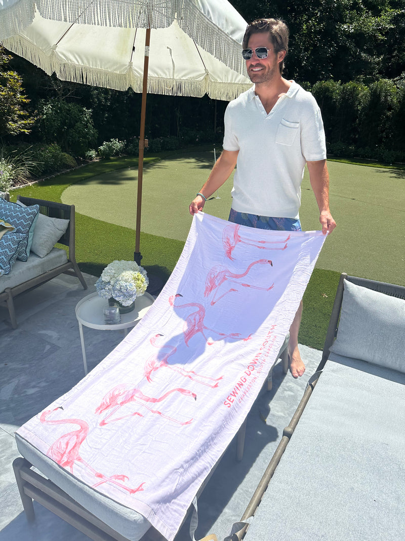 A man in sunglasses and a white shirt spreads a Flamingo Beach Towel on a poolside lounger beside a small table with flowers, under a white patio umbrella.