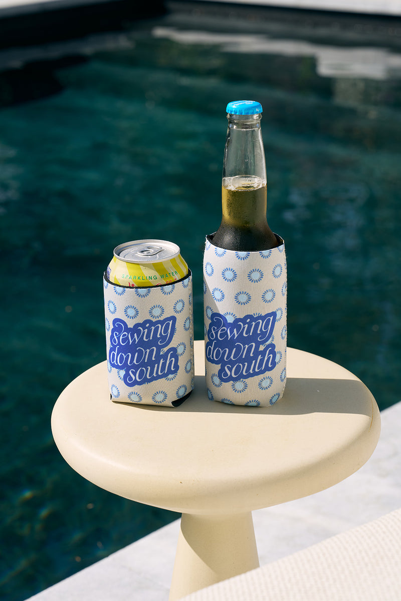Two drinks with SDS Calico Mini Floral Koozies sit on a small round table by the pool—a sparkling water can and a beer bottle. The background showcases clear blue water, each drink perfectly insulated by its stylish cooler.