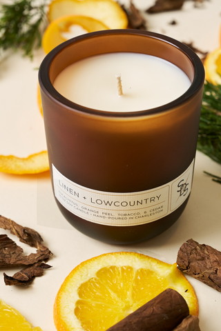 The Linen + Lowcountry Signature Candle, hand-poured in Charleston and set in a brown glass jar, sits unlit on a surface with orange slices, pine sprigs, and dried wood, offering a fresh and natural atmosphere.