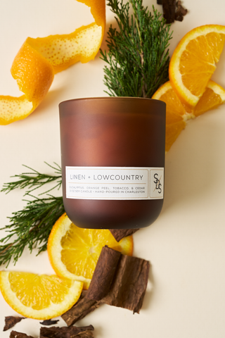 The Linen + Lowcountry Signature Candle, a brown glass soy candle hand-poured in Charleston, is displayed on a light background with orange slices, orange peels, cedar sprigs, and bark pieces.