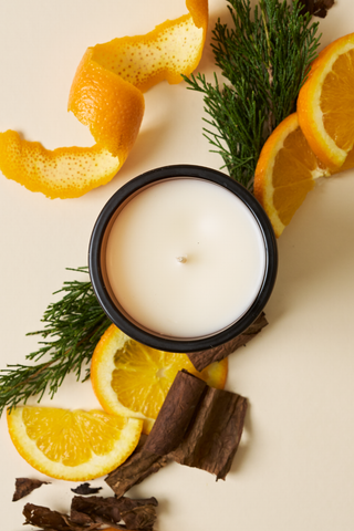 The Linen + Lowcountry Signature Candle, in a black container, is displayed on a light surface with orange slices, orange peel, pine branches, and pieces of brown bark arranged around it.