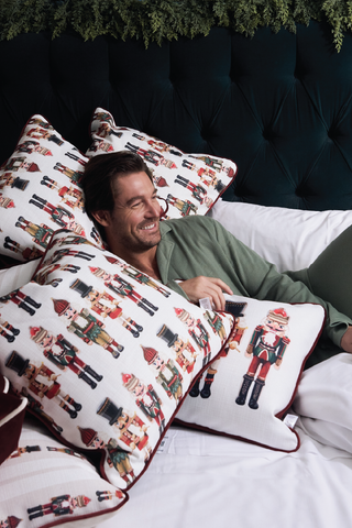 A man smiles while relaxing on a bed decorated with multiple Nutcracker Line Up Pillows and holiday pillows, holding a cup. Greenery hangs above the dark tufted headboard in the background.