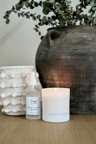 A large dark ceramic pot with green foliage sits on a wooden surface. In front, theres a knitted white container, a labeled SDS Signature Scent Room Spray infused with Jasmine Lily Patchouli Musk, and a lit white candle in a glass labeled Sewing Down South Candles.