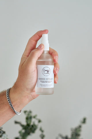 A hand holds a clear “Room Spray” bottle against a light background, with greenery below and a silver chain bracelet visible—create the perfect scent combo with the Mystery Candle + Room Spray Bundle.