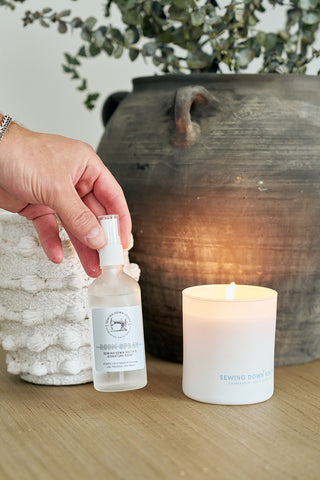 A hand holds the Mystery Candle + Room Spray Bundle—a spray beside a lit white soy candle on a wooden surface, with a rustic vase and greenery in the background.