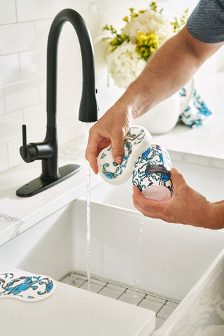 Using Crab Craze Pop Up Sponges, a person washes two patterned drinking glasses under a running tap in a white kitchen sink. The black faucet contrasts with the white flowers in the background, while the counter and backsplash remain pristine white.