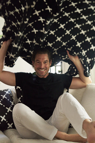 A smiling man in a black shirt and white pants sits on a couch, playfully lifting The Paige Collection: Exes in Black Blanket above his head. A cozy matching pillow with white X patterns rests beside him.