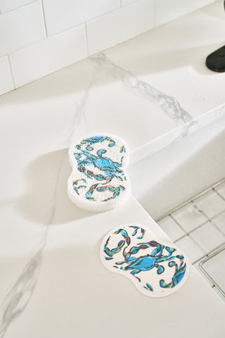 The Crab Craze Kitchen Towel Set is shown on a white marble countertop near a sink, alongside a pot holder, spatula, and blue-and-teal bird-designed pop up sponges. An oversized kitchen towel is folded neatly nearby.