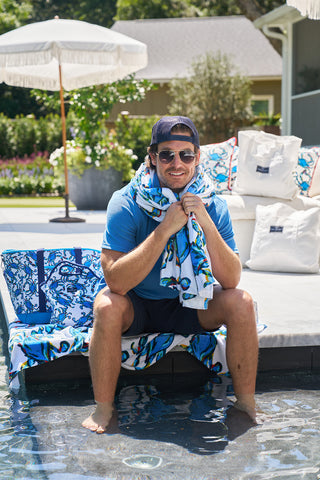 A man in sunglasses and a cap sits by the pool, smiling with a colorful Crab Craze Beach Towel from Sewing Down South draped around his shoulders. Hes sporting a blue shirt and shorts, with decorative pillows and an umbrella in the background.