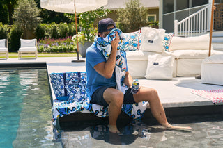 A man sits by the pool in a cap, blue shirt, and shorts. As he wipes his face with a towel while holding a can, the vibrant Crab Craze Beach Towel from Sewing Down South is next to him. White outdoor furniture sets the background.